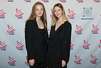 Vor einer weißen Wand, die mit mehreren SpaceNet Awards-Logos versehen ist, stehen zwei Frauen in schwarzer Kleidung