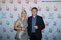 Vor einer weißen Wand mit mehreren SpaceNet Awards-Logos stehen ein Mann und eine Frau