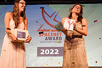 Katja Holzer steht mit einer Teilnehmerin des SpaceNet Awards vor einer Leinwand, auf der das SpaceNet Award-Logo abgebildet ist