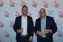 Vor einer weißen Wand mit mehreren SpaceNet Awards-Logos stehen zwei Männer in Anzügen