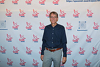 Vor einer weißen Wand mit mehreren SpaceNet Awards-Logos steht ein Mann in einem blauen Hemd