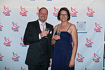 Vor einer weißen Wand mit mehreren SpaceNet Awards-Logos stehen ein Mann im Anzug und eine Frau im Kleid