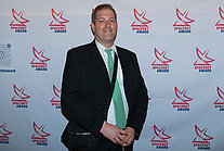 Ein Mann im Anzug mit grüner Krawatte steht vor einer weißen Wand, die mit mehreren Logos des SpaceNet Awards bedeckt ist