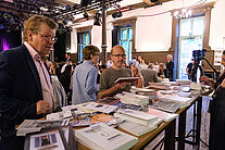 Zwei Männer stehen vor einem Tisch, der mit Büchern bedeckt ist, im Hintergrund sitzen mehrere Personen an Tischen