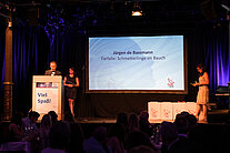 Drei Personen stehen auf der Bühne, eine davon steht vor dem Rednerpult und stellt einen Teilnehmer der SpaceNet Awards vor