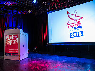 Ein Stehtisch auf einer Bühne, im Hintergrund auf einer Leinwand das SpaceNet Award Logo 2018