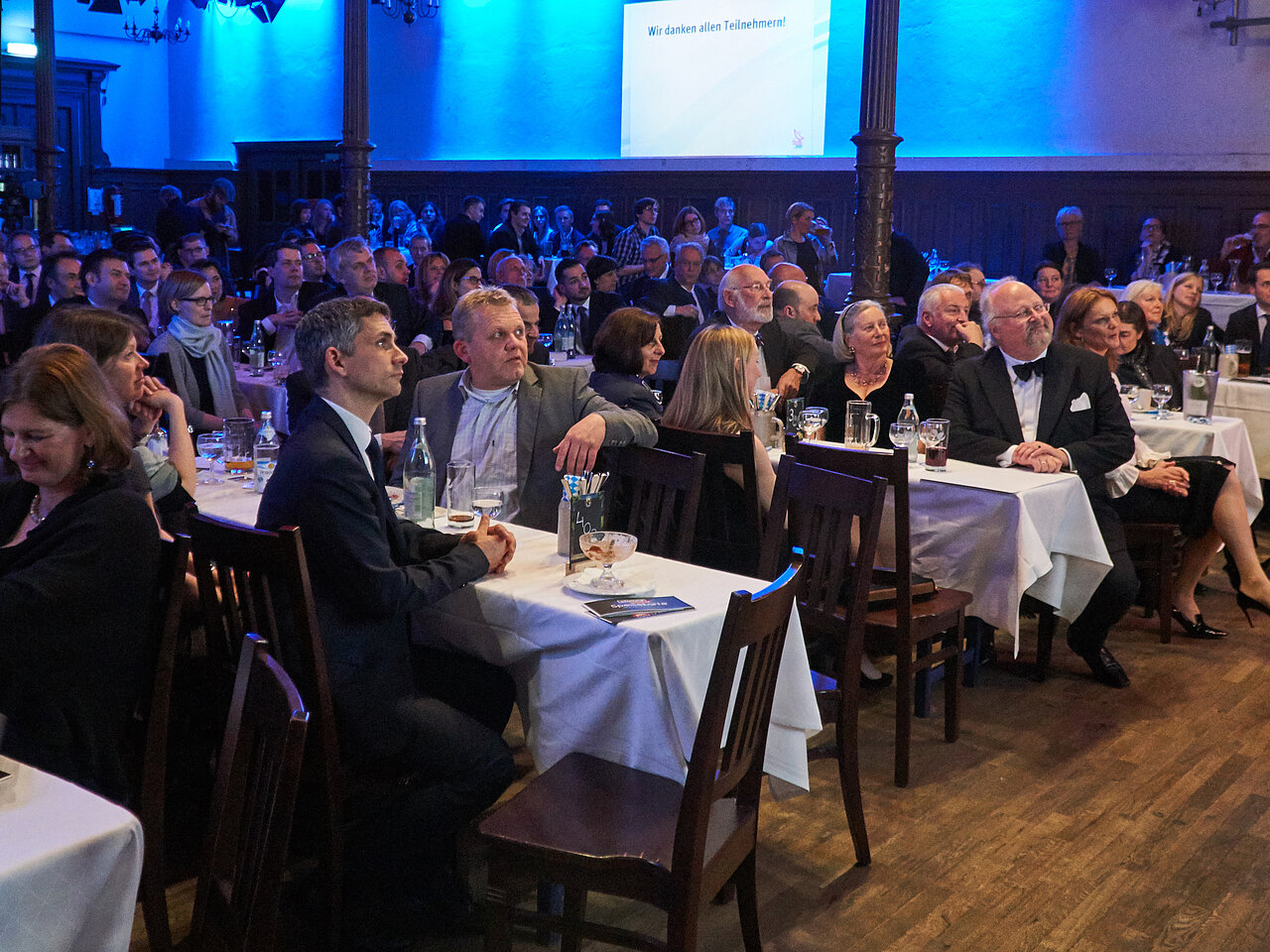 Mehrere Personen sitzen an den Tischen und hören aufmerksam einer Rede zu