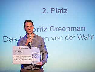 Ein Mann in einem grauen Hemd hält eine Rede auf einer Bühne und hält in seinen Händen ein Mikrofon sowie einen Preis