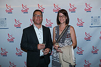 Vor einer weißen Wand mit mehreren SpaceNet Awards-Logos stehen ein Mann im Anzug und eine Frau im Kleid