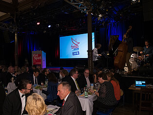 Mehrere Personen sitzen an Tischen und essen. Im Hintergrund sind das SpaceNet Award Logo und zwei Männer, die Instrumente spielen