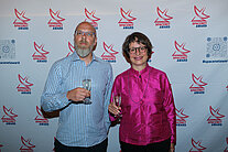 Ein Mann und eine Frau stehen mit einem Glas Champagner vor einer weißen Wand, die mit mehreren SpaceNet Award Logos bedeckt ist